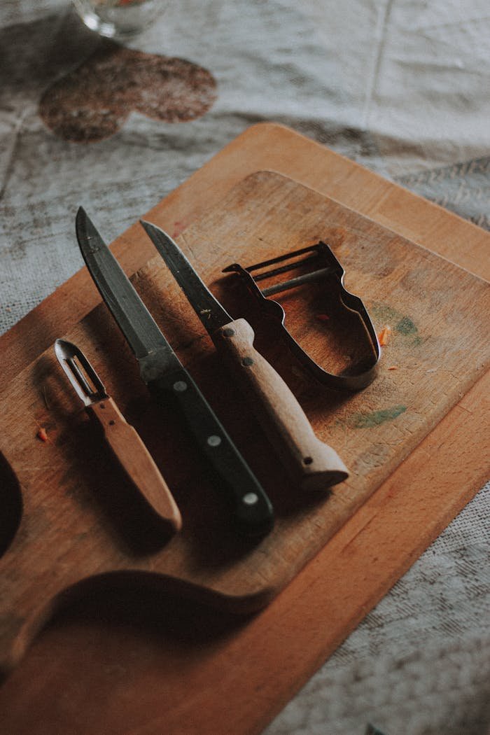 Close-up of knives and peelers on a wooden chopping board in a rustic kitchen setting.